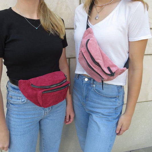 Pink corduroy belly bag around the shoulder of a girl wearing a white shirt and jeans next to a cream tile stone wallRed corduroy belly bag around the waist of a girl wearing a black shirt and jeans next to a cream tile stone wall and a 
